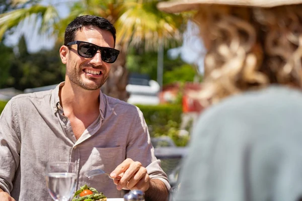 At Ondres Plage, a man grinning while eating outside beneath palm trees, grasping a fork.