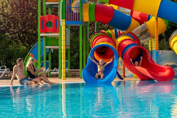 At Oliva, vibrant waterslides are being enjoyed by children as they plunge into a pool, while adults relax by the water's edge surrounded by lush foliage.