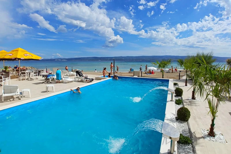 At Camp Galeb, a pool filled with individuals swimming and unwinding offers a view of a bustling beach and ocean beneath a clear sky adorned with scattered clouds.