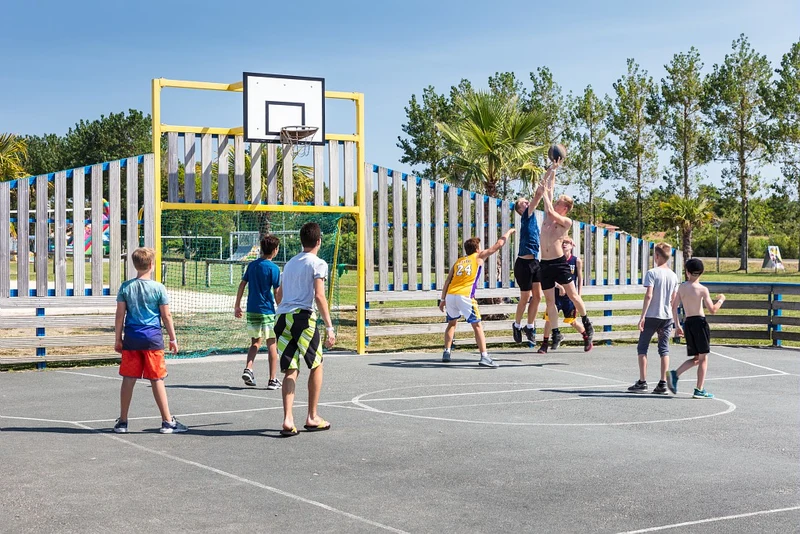 At Le Vieux Port, children are playing basketball on an outdoor court, with lush green trees and a clear blue sky in the background.
