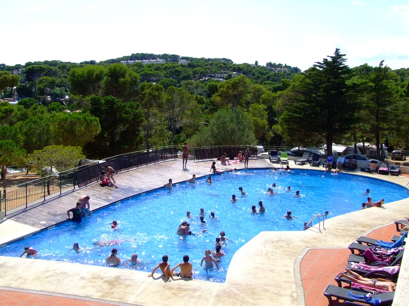 At Internacional de Calonge, a packed swimming pool features individuals swimming and relaxing, encircled by trees and a hilly terrain.