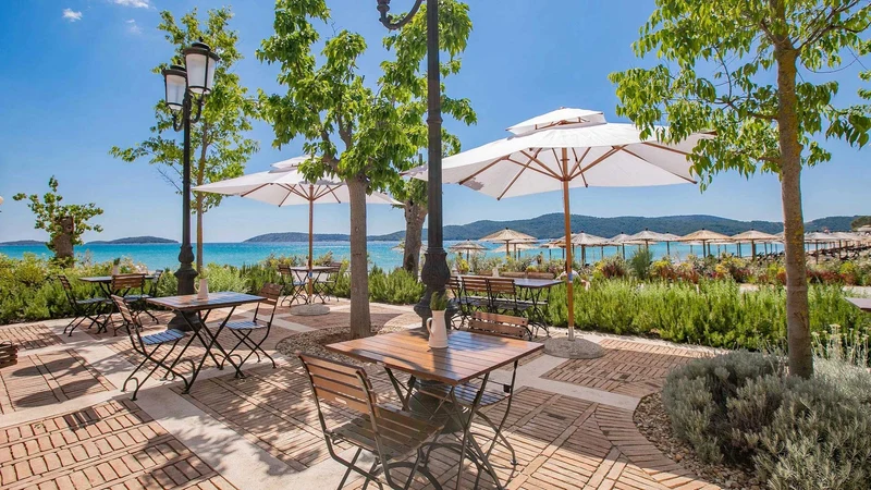 At Amadria Park Camping Sibenik, wooden tables and chairs are positioned beneath white umbrellas, providing a view of a picturesque beach surrounded by vibrant greenery and trees.