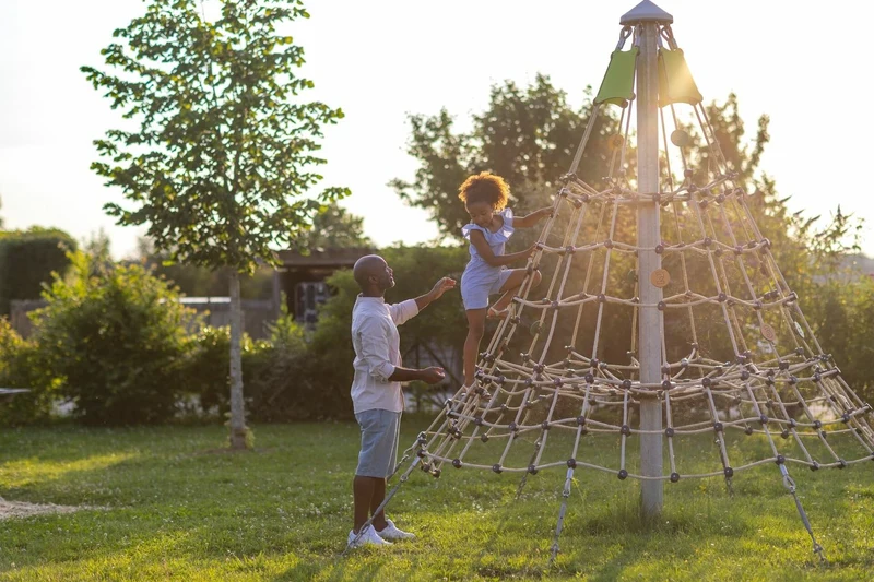 At Village Flottant de Pressac, a child ascends a rope pyramid, assisted by an adult, encircled by trees and lush vegetation.