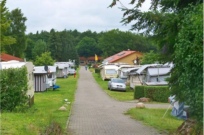 At Knaus Campingpark Bleckede/Elbtalaue, a surfaced walkway flanked by mobile homes and lush vegetation features an individual strolling in the mid-distance.