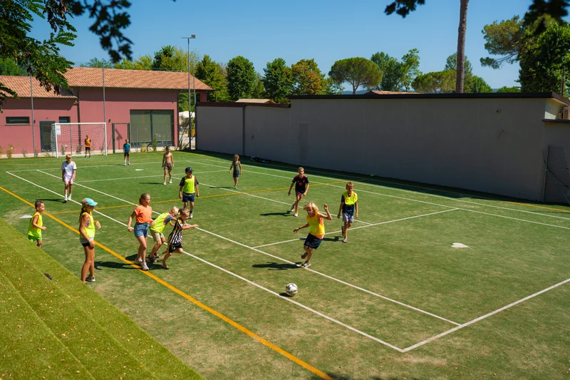 At Trasimeno Glamping Resort, kids are playing soccer on a compact artificial grass field encircled by structures and trees.