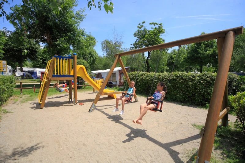 At Orchidea, kids play on swings and slides in a play area, encircled by sand and trees.