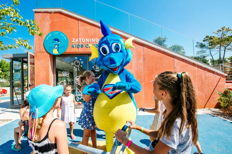 At Zaton Holiday Resort, a cheerful blue dragon mascot engages playfully with children outside a vibrant building on a sunny day.