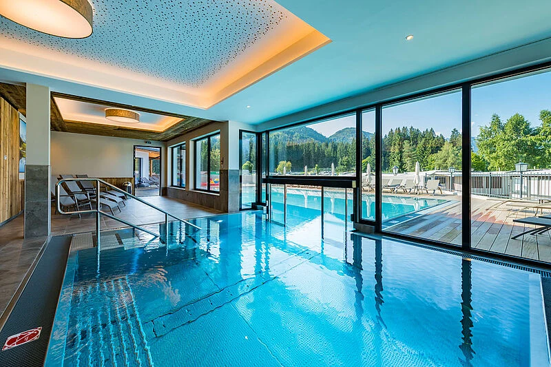 At Eurocamp Wilder Kaiser, an indoor pool seamlessly connects with an outdoor pool, framed by expansive windows that highlight stunning mountain vistas, accompanied by nearby lounging chairs.