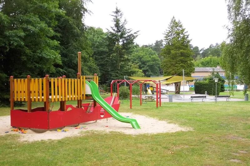 At Knaus Campingpark Bleckede/Elbtalaue, there is a children's play area featuring a slide and climbing equipment on grass, situated near a swimming pool and benches.