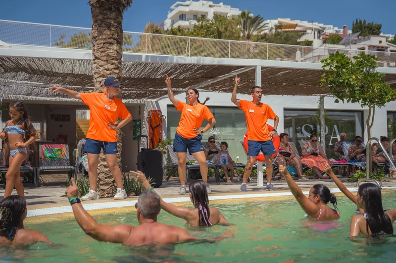 At Costa del Sol Glamping Village, instructors conduct a poolside workout session, gesturing animatedly, surrounded by swimmers and onlookers.
