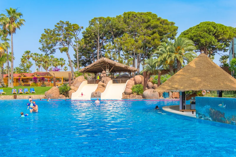 At Cambrils Park, kids are enjoying themselves in a large swimming pool featuring water slides, encircled by trees and sun loungers.