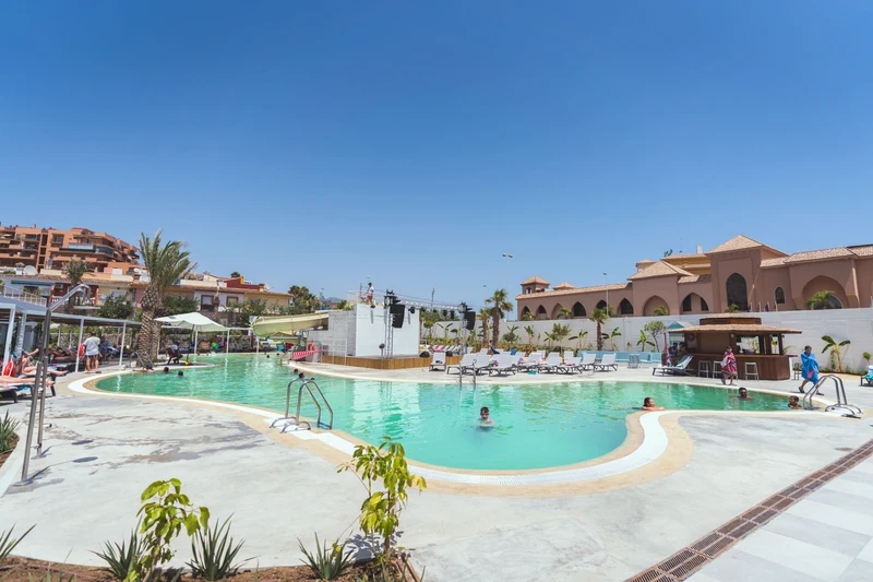 At Costa del Sol Glamping Village, a swimming pool features individuals unwinding beneath umbrellas, encircled by loungers and Mediterranean-style structures.