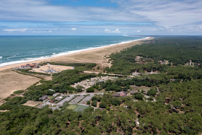At Ondres Plage, a coastal campsite is encircled by vibrant woodland and a sandy shore beneath a partly cloudy sky.