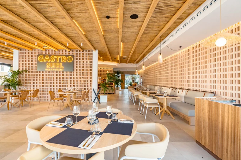 At Costa del Sol Glamping Village, tables are arranged for dining, encircled by light wooden decor and greenery, in a contemporary restaurant atmosphere.