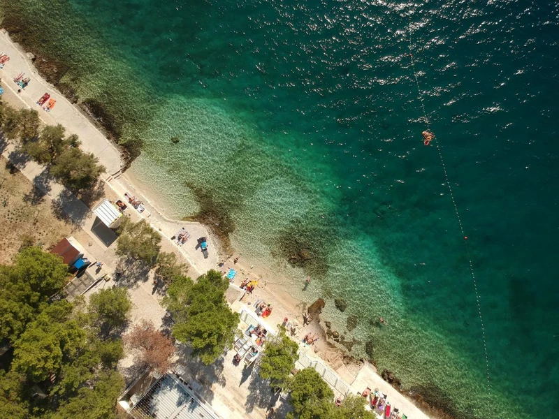 At Amadria Park Camping Trogir, vacationers are unwinding on sunbeds along a tree-fringed coastline with crystal-clear turquoise waters.